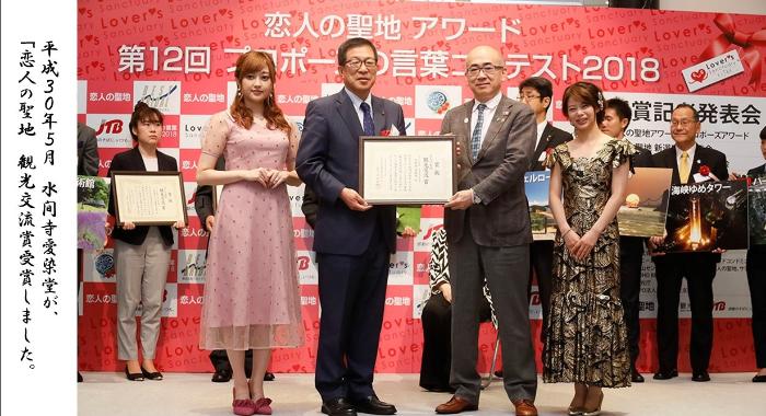 平成30年5月 水間寺愛染堂が恋人の聖地 観光交流賞を受賞しました。「恋人の聖地アワード 第12回プロポーズの言葉コンテスト2018」と書かれた背景の前で、額縁に入った賞状を手に持って記念撮影をしています。