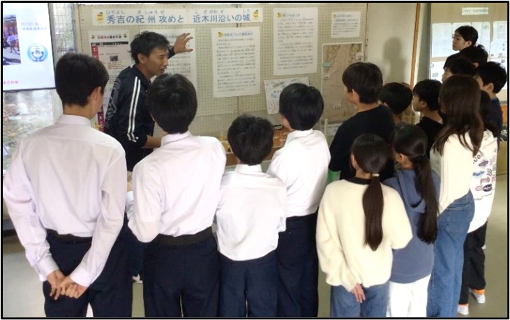 二色学園の玄関付近で小学生・中学生がともに展示の解説を聞くようす