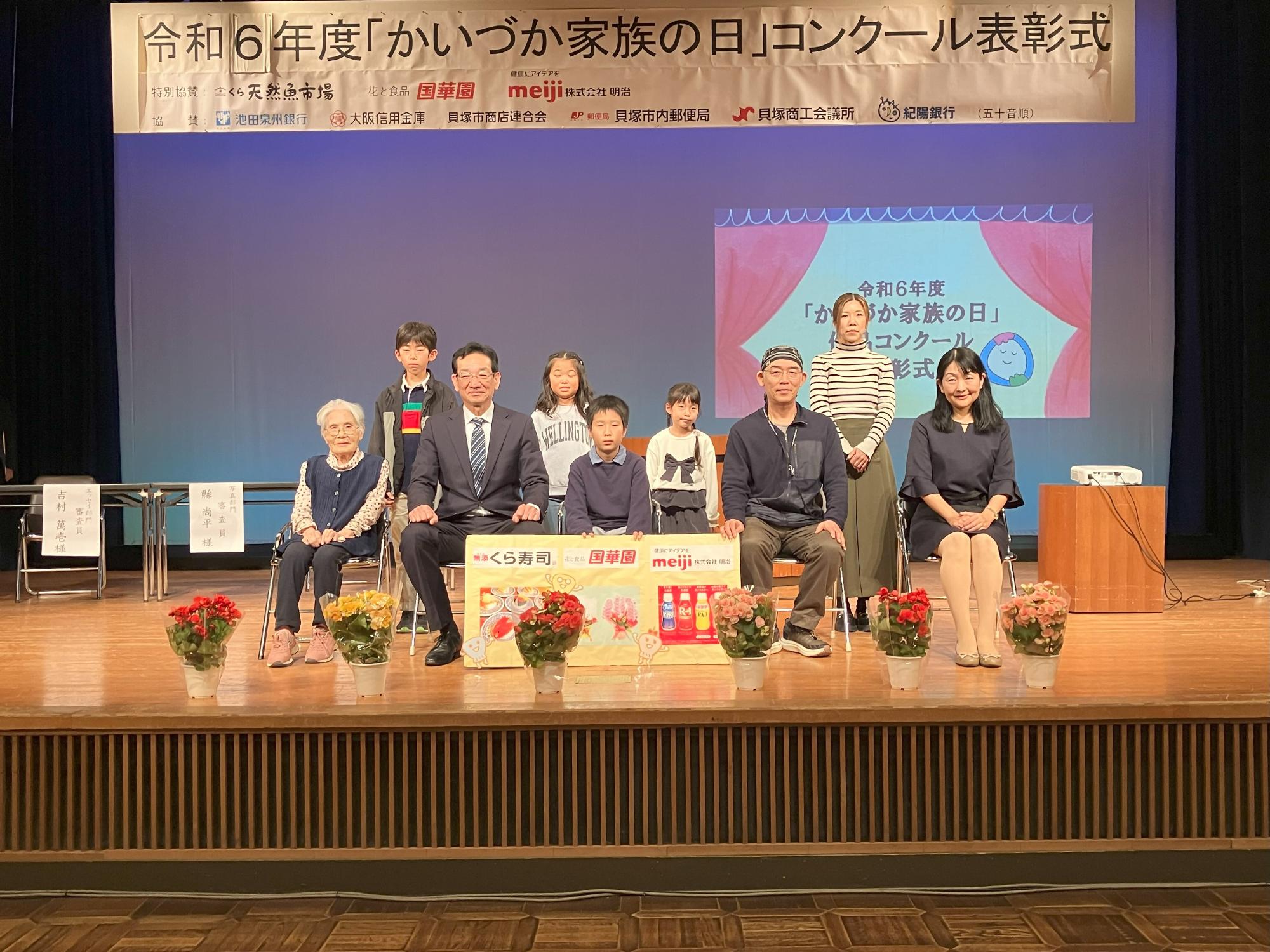 エッセイ部門記念撮影  特別協賛のくら寿司、国華園、meiji(明治)のパネルとともに集合写真を撮影しました(舞台上のスクリーンに『令和5年度「かいづか家族の日」作品コンクール表彰式』のスライドが映し出されている)