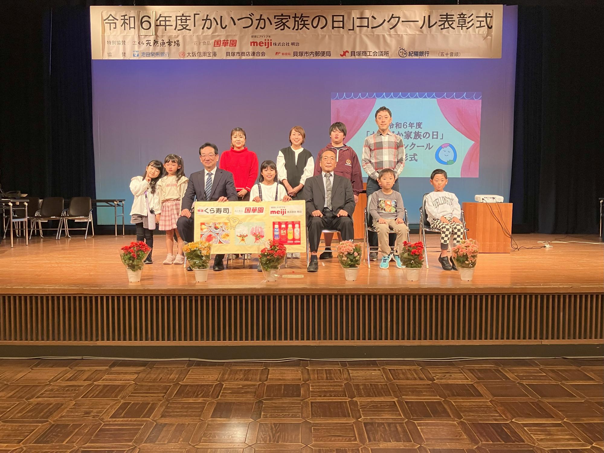 写真部門記念撮影  特別協賛のくら寿司、国華園、meiji(明治)のパネルとともに集合写真を撮影しました(舞台上のスクリーンに『令和5年度「かいづか家族の日」作品コンクール表彰式』のスライドが映し出されている)