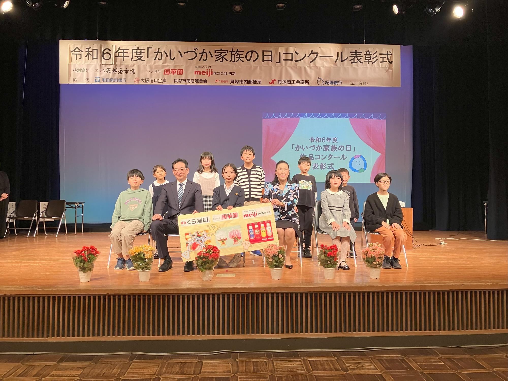絵画部門記念撮影  特別協賛のくら寿司、国華園、meiji(明治)のパネルとともに集合写真を撮影しました(舞台上のスクリーンに『令和5年度「かいづか家族の日」作品コンクール表彰式』のスライドが映し出されている)