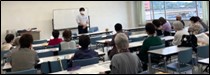 講演会の参加者の皆さん(立って話をする講師の話を、10名以上の参加者が各々着席して聞いている様子)