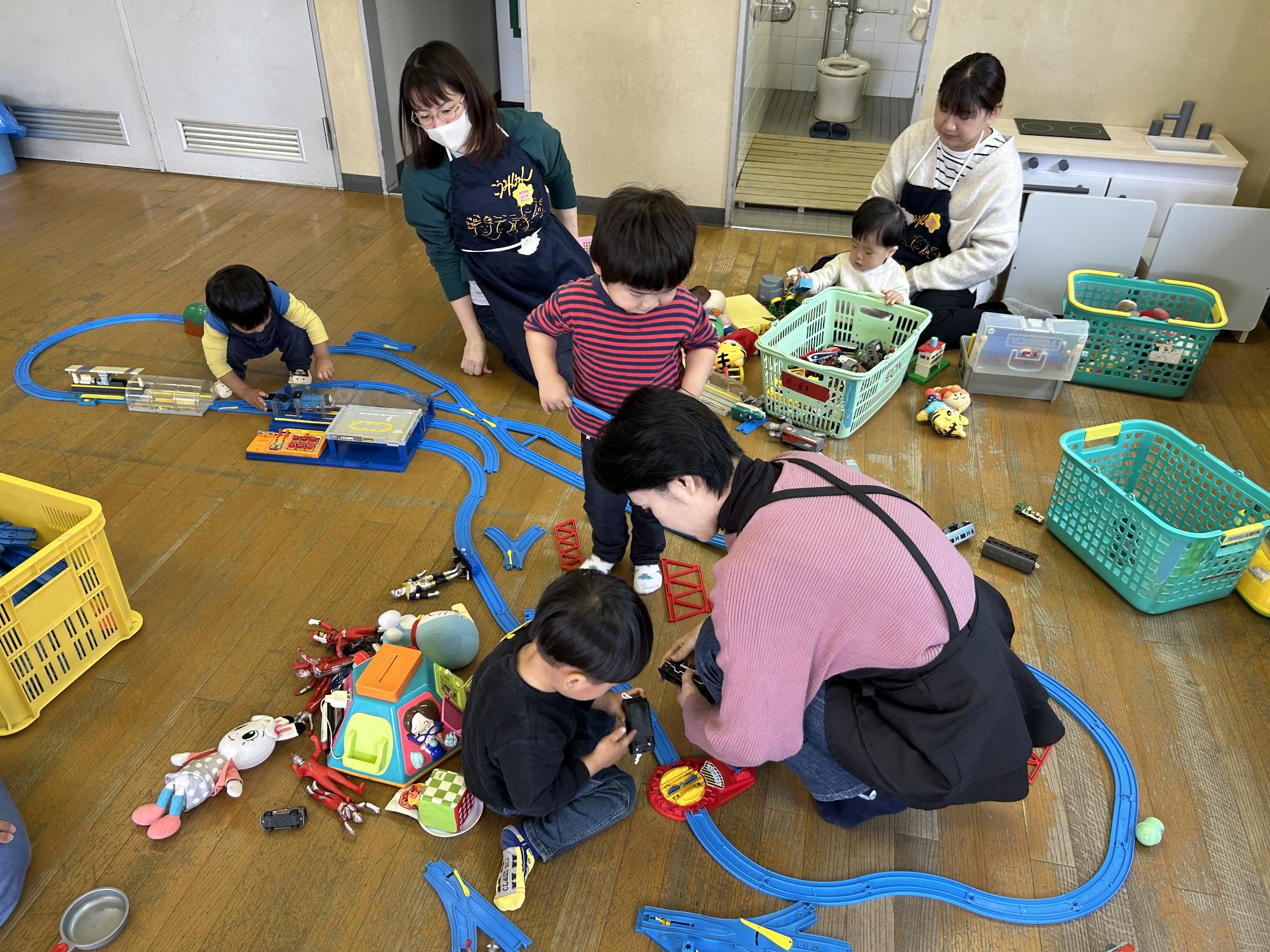 保育室で乳幼児のこども4人が電車やおもちゃを広げて遊び、保育ボランティアが子どもの遊びを見守っている
