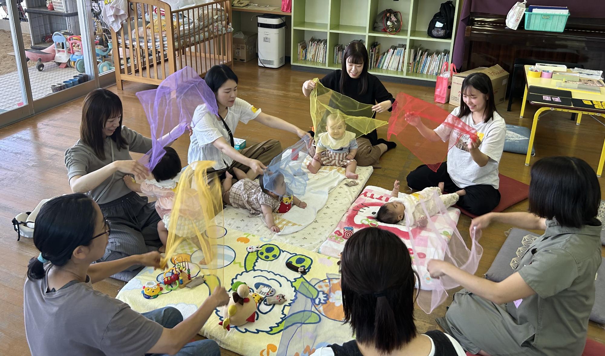 布団の上に赤ちゃんたちが寝転がっていて、お母さんたちが色とりどりのオーガンジー布で赤ちゃんと笑顔でふれあっている