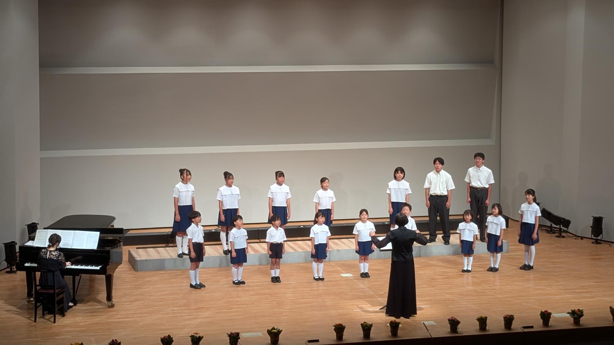 大きいホールでの舞台に制服を着た子どもたちが2列に並び、指揮者やピアノに合わせて合唱している