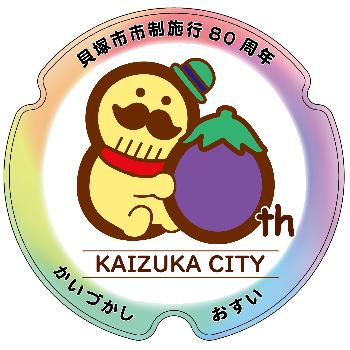 市制80周年記念ロゴマークがデザインされたマンホール蓋(市の伝統工芸品のつげ櫛がモチーフのイメージキャラクターの「つげさん」と特産品の水ナスで「80」を見立たデザイン。マンホールデザインの周りには虹色の縁あり)