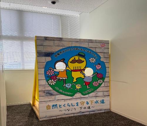 市役所本館2階山側トイレ横に設置された、市制80周年記念マンホールがデザインされた顔出しパネル(つげさんが子ども2人と手を繋いでおり、子ども2人の顔が顔出しできるようになっている)