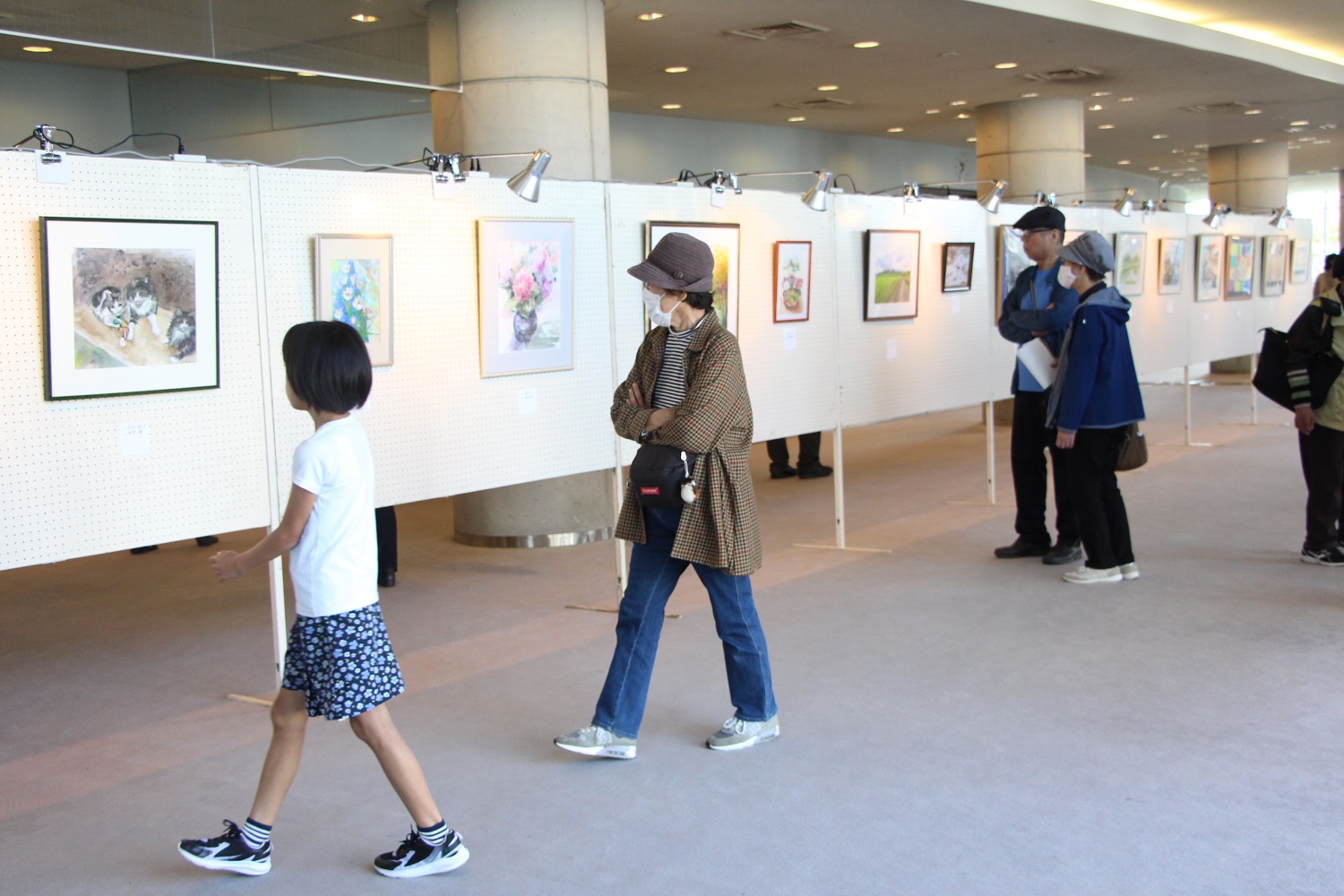 複数のパネルに絵画などの作品を展示し、来場者が見学している写真です。