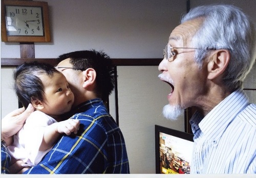 令和元年度かいづか家族の日の写真部門優秀賞作品です 父に抱っこされた生後2か月の赤ちゃんを祖父が大きな口を開けてあやしている様子を写した写真です 赤ちゃんはじっと祖父の顔を見つめています