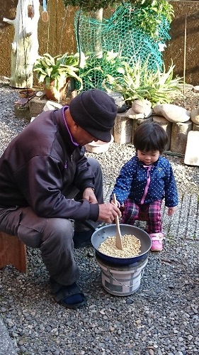 令和2年度かいづか家族の日の写真部門優秀賞作品です 屋外で祖父が七輪を使って大豆を炒っている様子を孫が見守っている様子を写した写真です