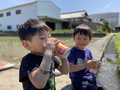 令和4年度かいづか家族の日の写真部門優秀賞作品です 青空の下 二人の男の子が田んぼのあぜ道に座ってジュースを飲んでいる様子を写した写真です ジュースを持つ手は腕まで泥がついています