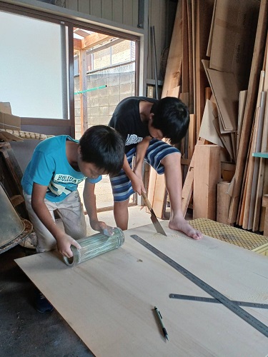 令和4年度かいづか家族の日の写真部門優秀賞作品です 作業場で 兄はのこぎりで板を切ってグローブ入れを作っており 弟は筒状のものをやすりでこすって貯金箱を作っている様子を写した写真です