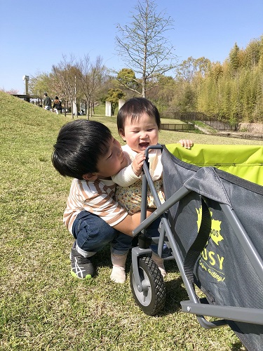令和4年度かいづか家族の日の写真部門優秀賞作品です 青空の公園の芝生の上で キャリーワゴンにつかまって立っている1歳くらいの妹と 後ろから抱きしめる7つ上の兄を写した写真です 兄は笑顔の妹をのぞきこんでいます