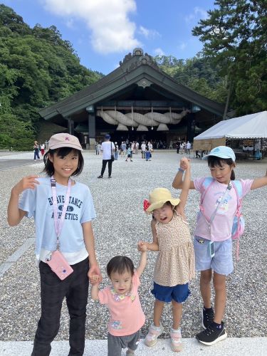 出雲大社を背景に幼児から小学生までの4姉妹が手を繋いでばんざいをして微笑んでいるようすを写した写真です。