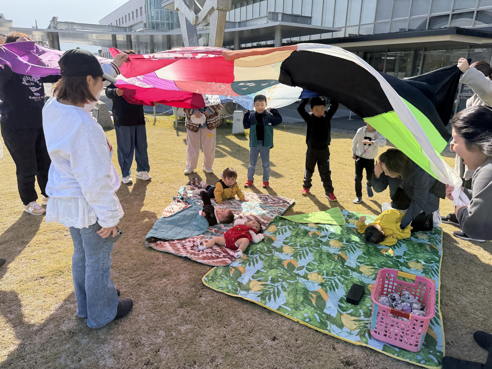 緑の芝生の上でカーペットをしき、赤ちゃんが寝ていて、お母さんや大きい子どもたちが大きな布をみんなでもって風で膨らみ、バルーン遊びを楽しんでいる様子