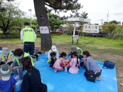 中央公民館の遊び隊（屋外の広場でブルーシートを敷き、遊び隊と子どもたちが輪になって座り遊んでいる）