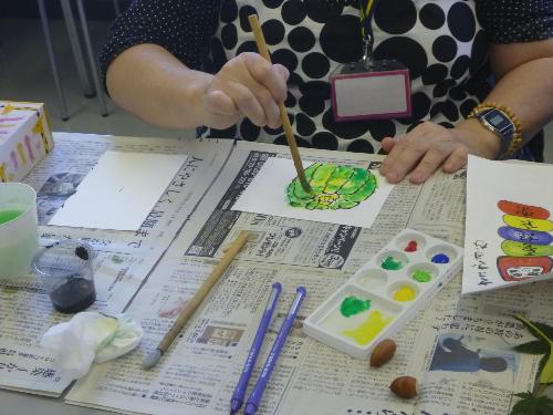机の上に新聞紙や絵の具セットを広げ、机に向かって筆を持ち、絵手紙に挑戦している様子