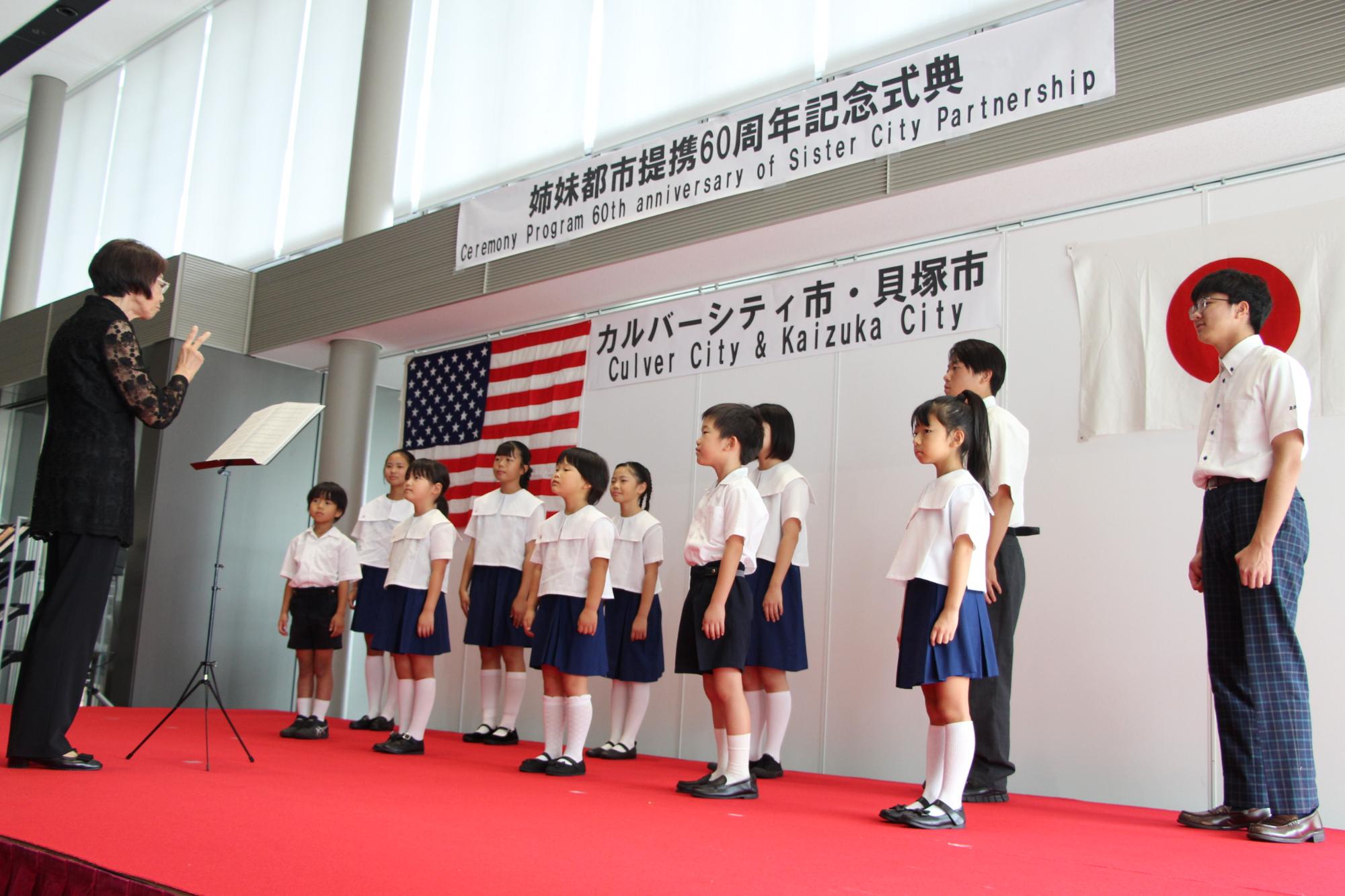 市役所ロビーで行われたアメリカのカルバーシティ市との姉妹都市提携60周年式典にて赤い舞台の上で制服を着た子どもたちが指揮者に合わせて歌っている