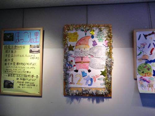 ほっこりの里の作品（12月  サンタクロースのイラストが画用紙いっぱいに描かれている  金や銀のモールで飾り付けされている）