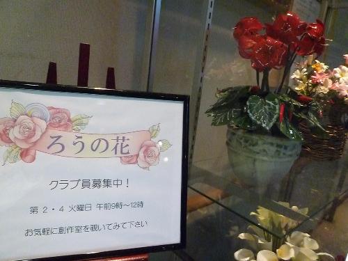 ろうの花（「クラブ員募集中  第2、第4火曜日午前9時～12時  お気軽に創作室を覗いてみてください」と書いた看板の隣に赤色の花が飾られている）
