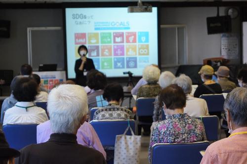 SDGs（エスディージーズ）について学ぶ様子です  会場前方のスクリーンには、「SDGsってなぁに？」の文字と17の目標のアイコンが写っています