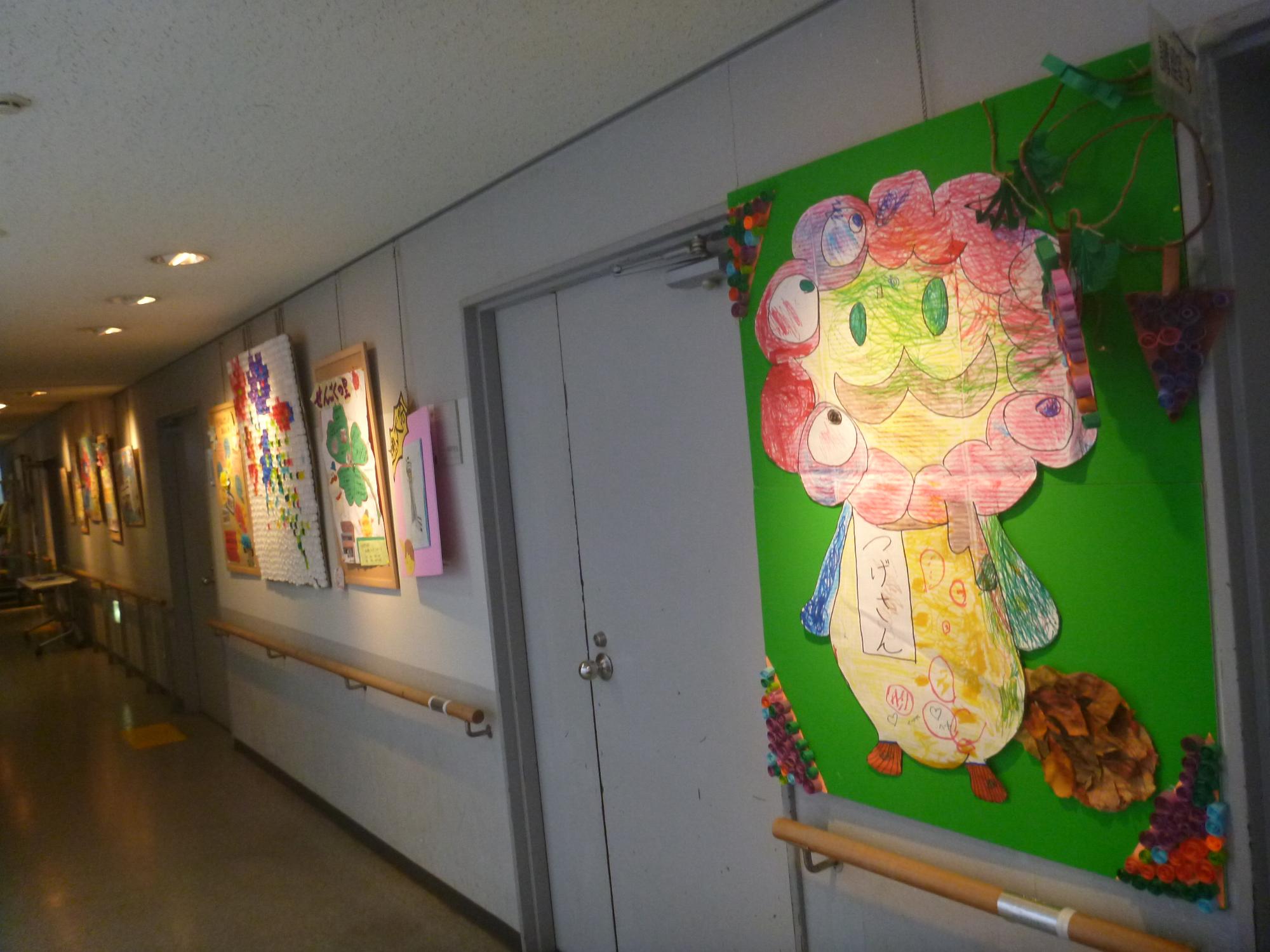 公民館のロビーに展示作品がずらっと展示されている 手前の作品は貝塚のキャラクターのつげさんと万博キャラクターのコラボされたイラストに色を塗ったり、ペットボトルのキャップで装飾された展示品などが飾られている