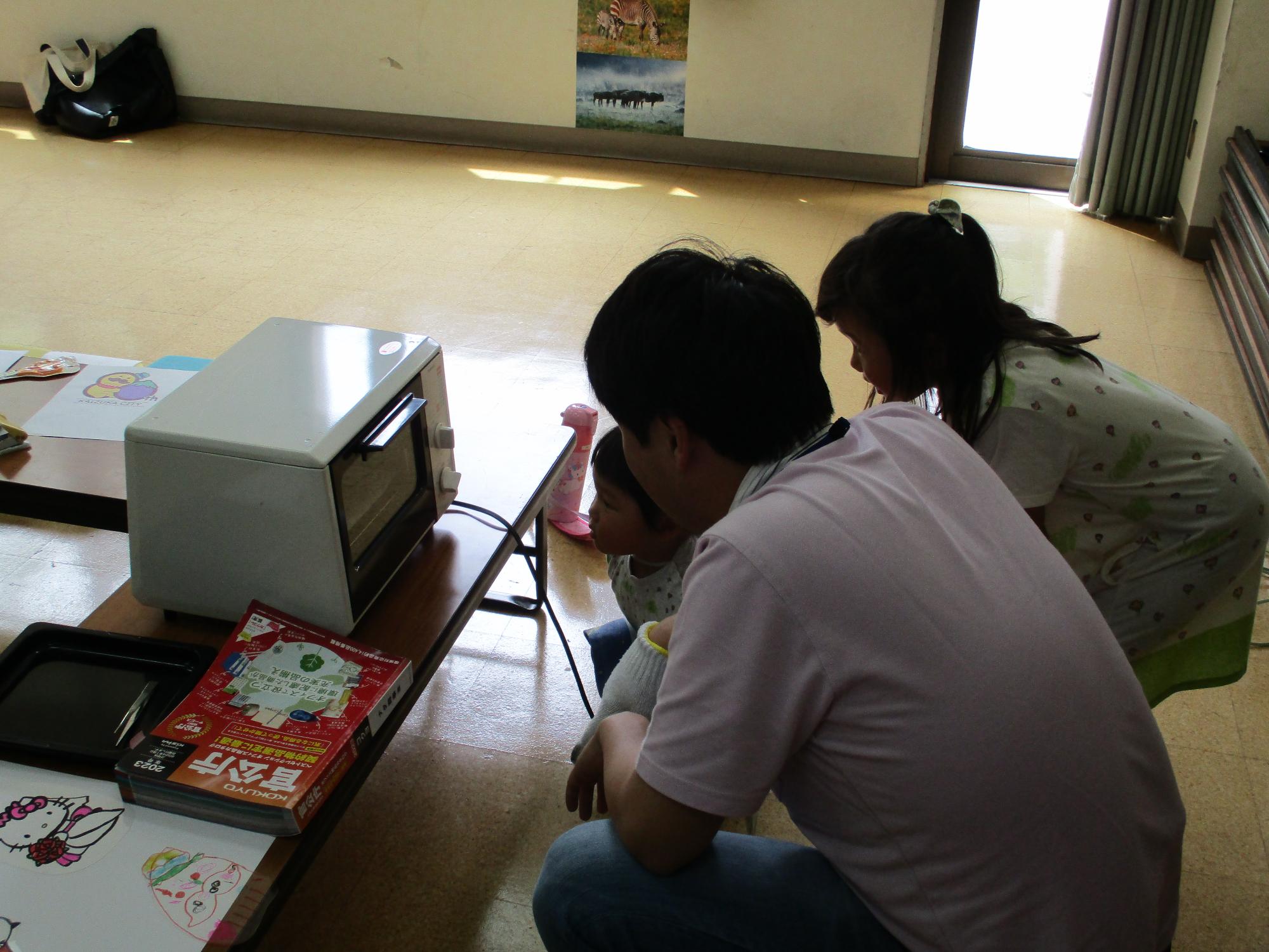 お父さんと子ども達が、プラ板が焼けるのをオーブントースターの前で見ています。