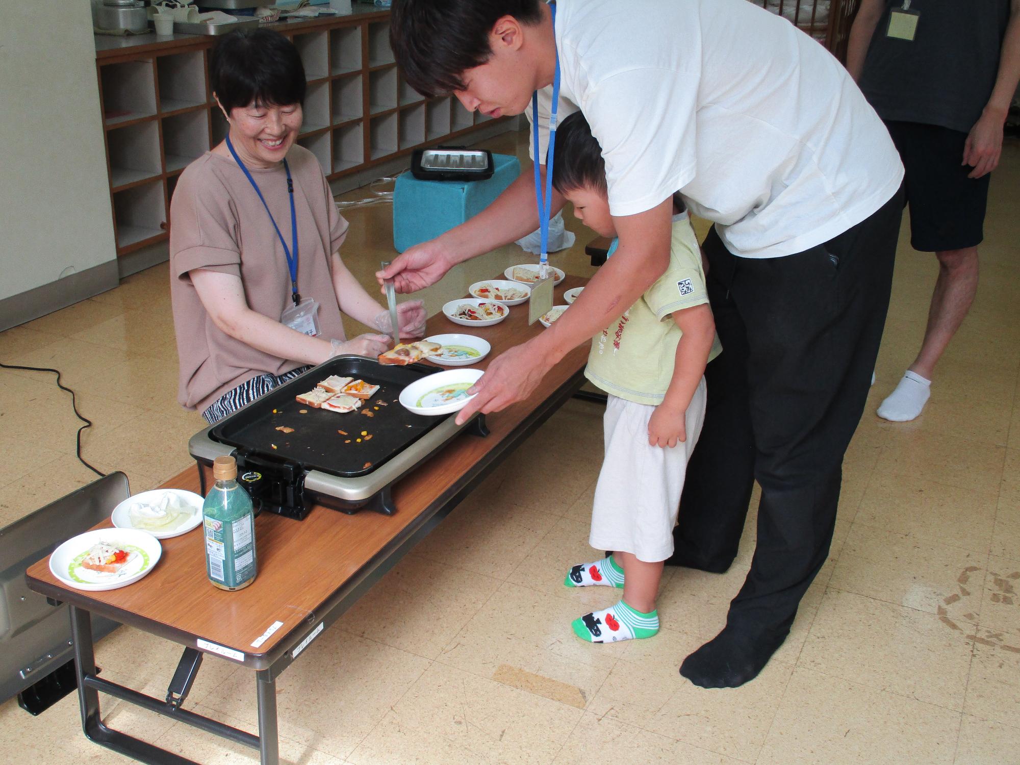 お父さんと子どもがホットプレートで焼いた食パンピザをお皿に取ろうとしています。