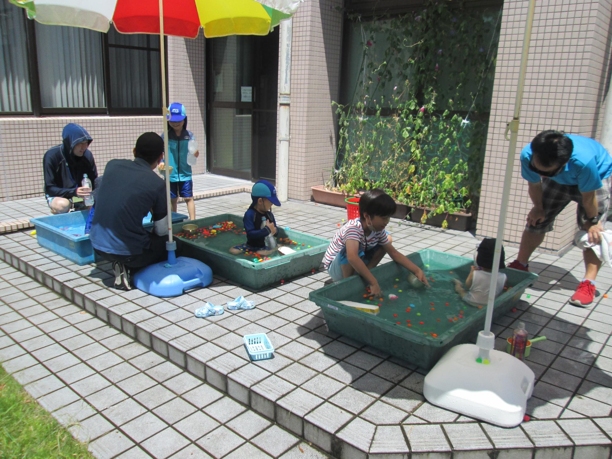 子ども達はプールの中に入り遊び、お父さんたちはプールの横で水遊びをしています。