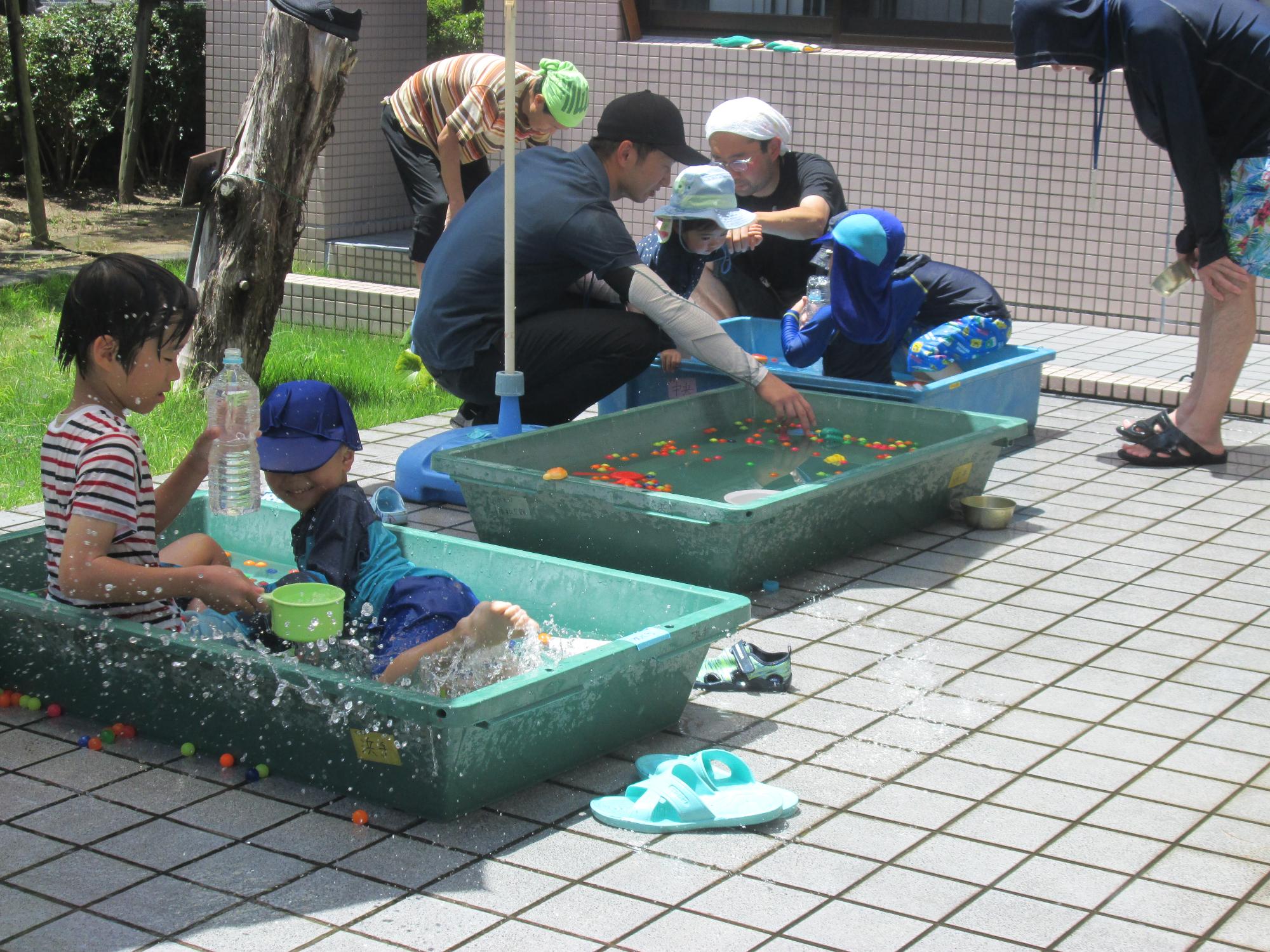 プールに入りペットボトルで水あそびをする子ども達と隣のプールで水あそびをしていいる親子。