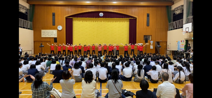 二色学園とのコラボで開催した「芸術鑑賞会」でストレッチ体操浜風のクラブ員達が出演（演者たちが両手を伸ばして上げている。  座っている二色学園の子ども達も同じように両手を伸ばして上げている。子ども達の後ろには保護者や地域住民も座っている。）