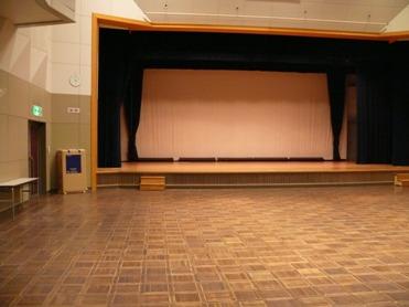 ホールの写真（正面に舞台  向かって左手に司会者台、長机が置かれている）