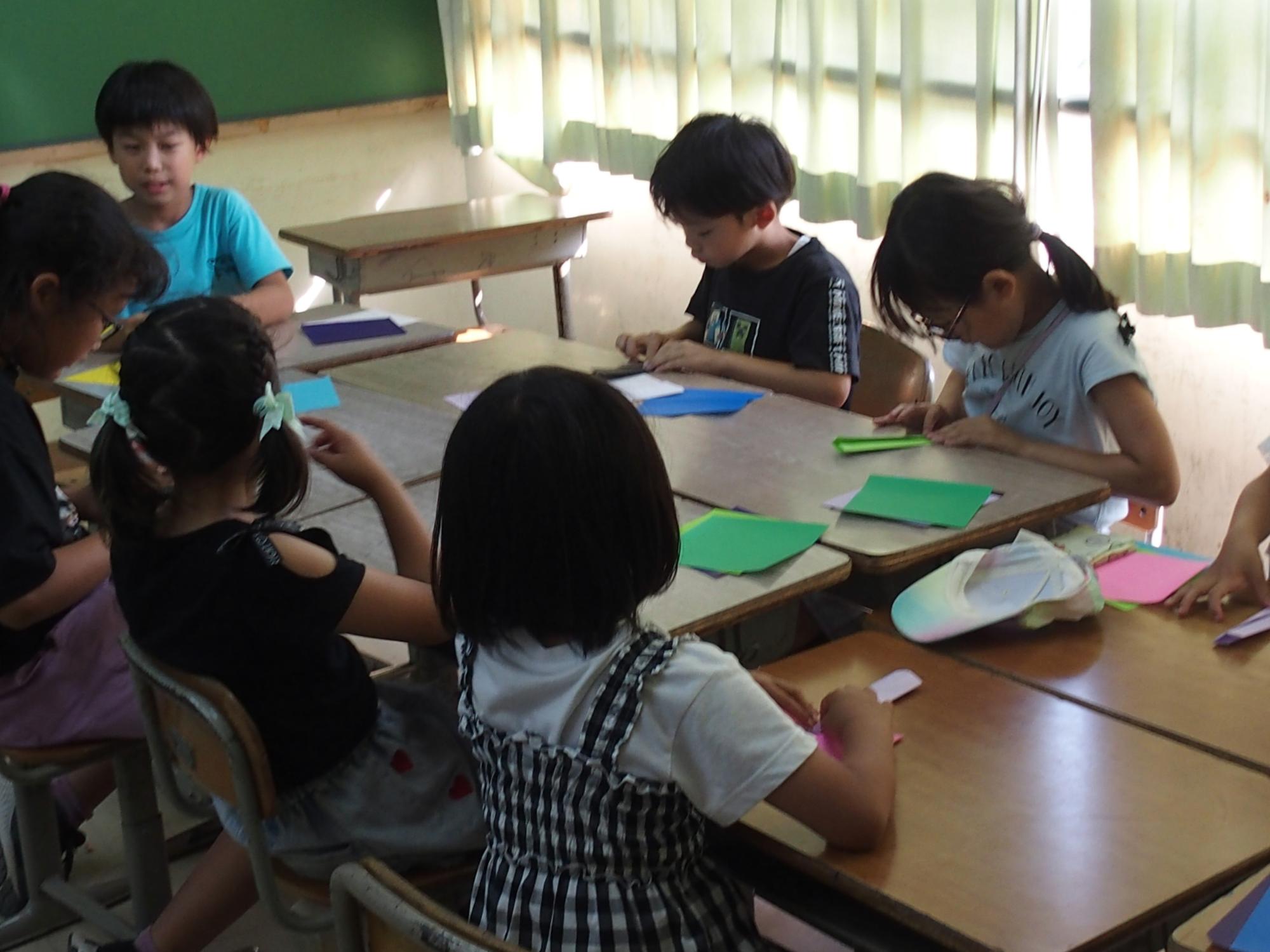 おりがみ活動（子どもたちが机を向かい合わせにひっつけて折り紙を折っています。）