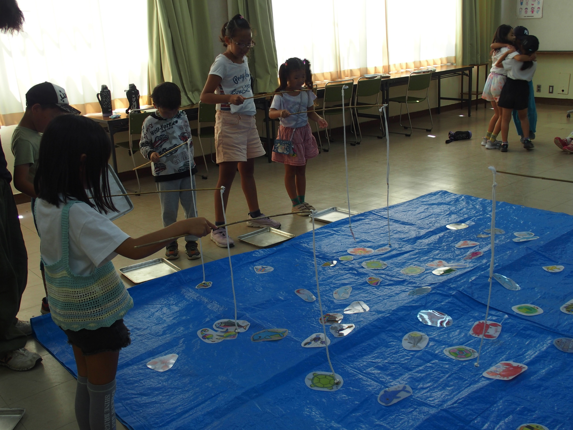 室内で子どもたちがブルーシートの上に並べられた手作りのさかなをつっている様子