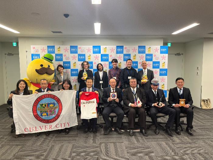 カルバーシティ市の高校生たちが、貝塚市長や貝塚市イメージキャラクター「つげさん」と集合写真を撮影しました。（CALIFORNIAとプリントされたTシャツやCULVER CITYの横断幕を手に持っている）