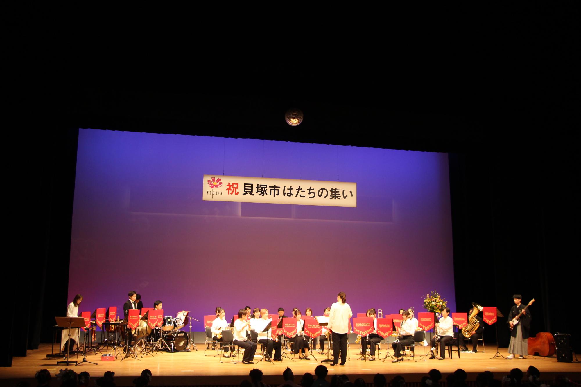 コスモスシアター大ホール舞台上にて市吹奏楽団がお祝い演奏をしている様子