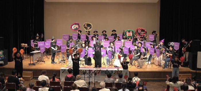 山手地区公民館の舞台上で楽器を演奏する団員や踊りをする団員たちがいて全員が楽しんでいる様子。