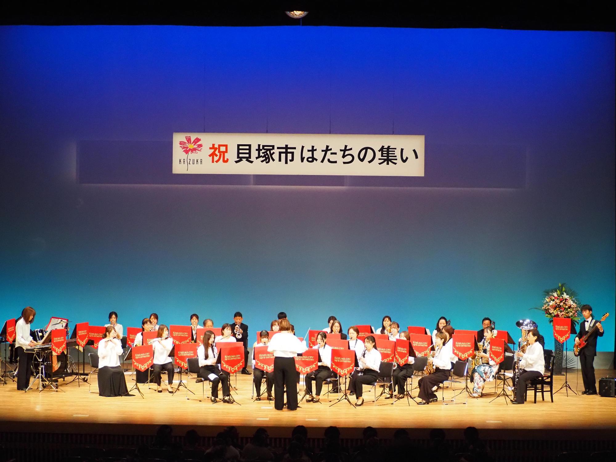 コスモスシアター大ホール舞台上にて市吹奏楽団がお祝い演奏をしている様子