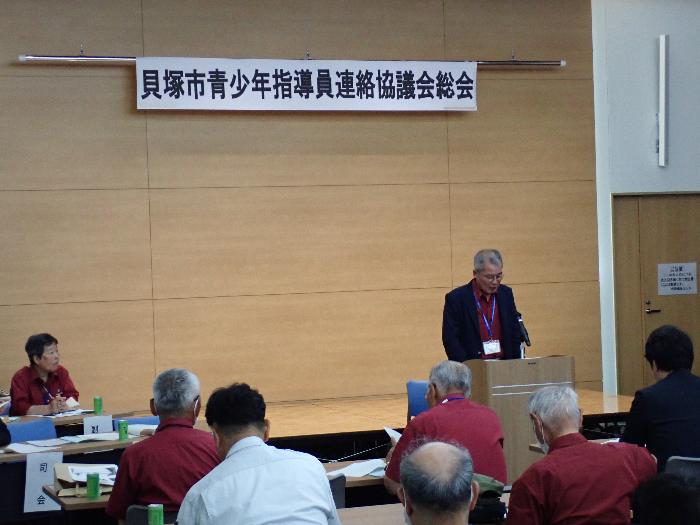 市役所内会議室にて総会時、えんじ色のTシャツを着用した青少年指導員たち集まり、そうの前で会長があいさつをしている様子