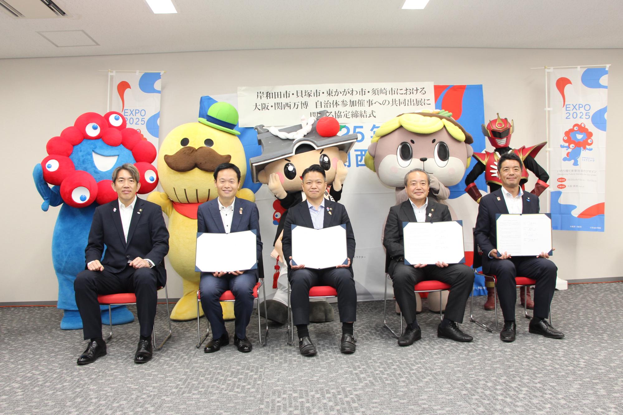 協定式の集合写真（前段に各市長(左から松原市長、貝塚市長、岸和田市長、須崎市長、東かがわ市長〉、後段にイメージキャラクター〈ミャクミャク、つげさん、ちきりくん、しんじょう君、てぶくろマン〉）