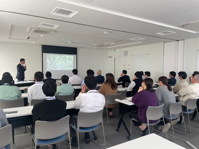 市職員を対象に、関西自動車学院の講師が座学講義をしている様子。会場前方のスクリーンには、講義内容のスライドが映し出されています。